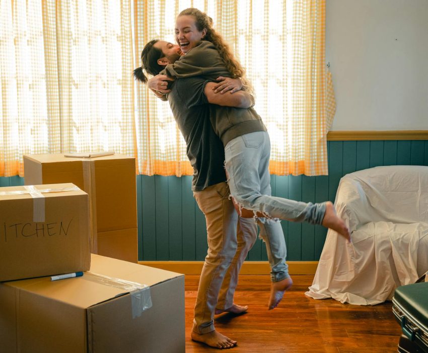 Side view full body barefoot young bearded male in casual clothes standing against window and lifting laughing girlfriend up during relocation and unpacking things from cardboard boxes
